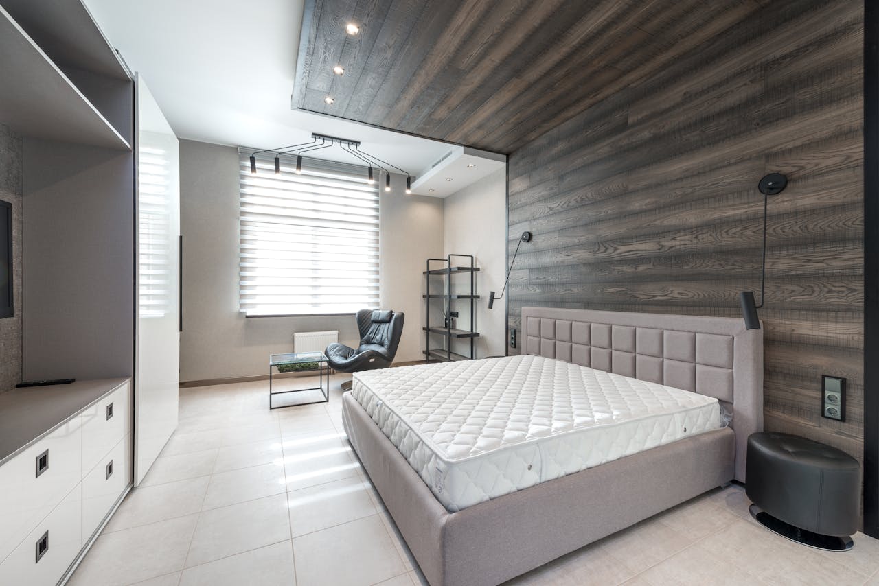 Bed with white mattress placed at wall with decorative element against cupboards in light stylish bedroom with window and armchair